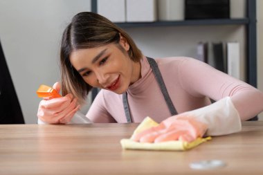 Eldiven ve önlük giymiş bir kadın, modern bir ev ortamında tahta bir yüzeyi sprey şişeyle ve bezle temizliyor..