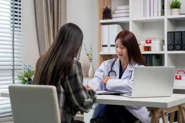 Beyaz önlüklü kadın doktor, modern ofis ortamında dizüstü bilgisayarı ve tıbbi ekipmanı olan bir hastaya danışıyor..