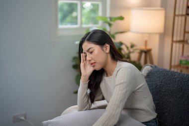 Evdeki bir kanepede oturan genç bir kadın, acı içinde başını tutuyor, baş ağrısı ya da stres tasvir ediyor..