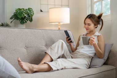 Rahat bir Asyalı kadın kanepede oturuyor, kahve içiyor, ve rahat bir oturma odasında kapalı salonda bitkilerle birlikte akıllı telefon kullanıyor.