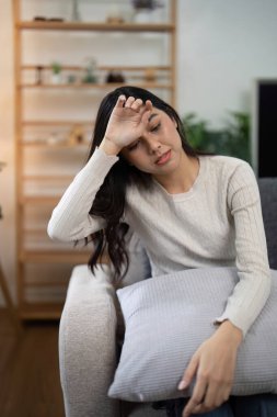 Kanepede oturan genç bir kadın, hasta ve yorgun görünüyor, elinde yastıkla. Görüntü rahatsız edici bir anı ve dinlenme ihtiyacını yakalar..