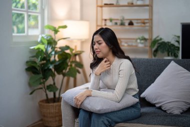 Evdeki bir kanepede oturan genç bir kadın, elinde yastıkla rahatsızlık yaşıyor, sağlık ve sağlık sorunlarını vurguluyor..