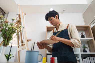 Önlük giyen genç bir sanatçı, doğal ışık ve çeşitli sanat malzemeleriyle dolu modern bir stüdyoda resim yapmaya odaklanmış..