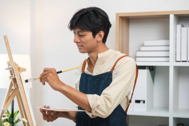 Önlük giyen genç bir erkek sanatçı, modern bir stüdyoda tuvale resim yapıyor ve arka planda raflar ve düzenli bir depo var..