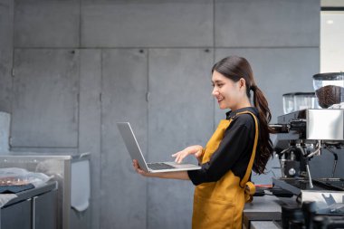Sarı önlüklü genç bir bayan barista modern bir kahve dükkanında dizüstü bilgisayar kullanıyor, gülümsüyor ve işiyle meşgul oluyor..