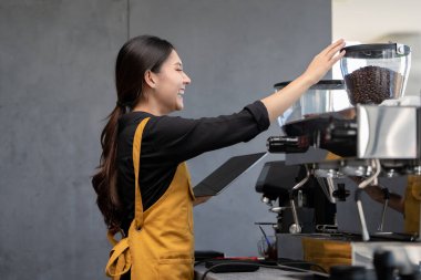 Sarı önlüklü gülümseyen barista modern bir kafede kahve öğütücüsü kullanıyor, elinde dijital tablet var..