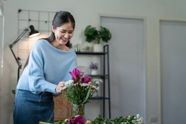 Mavi kazaklı bir kadın, doğal ışık ve yeşil bitkilerle dolu modern bir evde vazoda çiçekler düzenliyor..