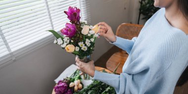 Mavi kazaklı bir kadın, pencerenin yanındaki vazoda bir buket taze çiçek düzenler, rahat ve huzurlu bir ev atmosferi yaratır..