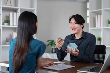 Çağdaş ofis ortamında müşteriyle yakınlaşan, ev modelini ve anahtarları sergileyen emlakçı..