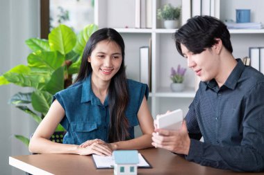 Emlakçı, modern ofis ortamında potansiyel bir alıcıyla mülk detaylarını tartışıyor. Minyatür bir ev modeli sergiliyor..