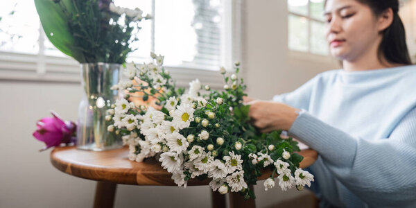 A woman in a light blue sweater arranges fresh white flowers in a vase on a wooden table in a bright room with natural light.