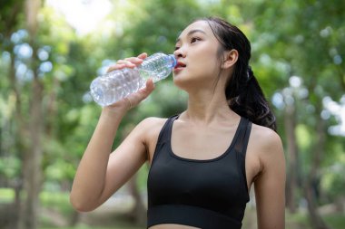 Sporcu genç bir kadın yemyeşil bir parkta egzersiz yaparken şişeden su içiyor..