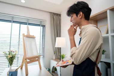 Modern bir stüdyoda genç bir sanatçı, düşünceli bir şekilde sehpadaki boş bir tuvale bakıyor, doğal ışık ve yaratıcı aletlerle çevrili..