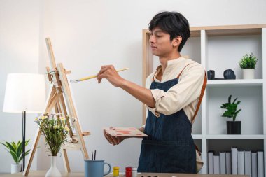 Modern bir stüdyoda, arka planda bitkiler ve raflar olan bir tuvalde resim yapan genç bir sanatçı..