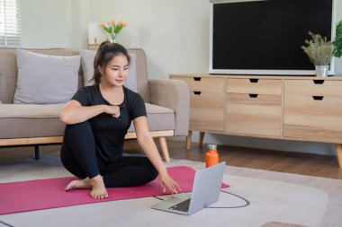 Evde yoga yapan bir kadın, rahat bir oturma odasında online fitness dersi için dizüstü bilgisayar kullanıyor..