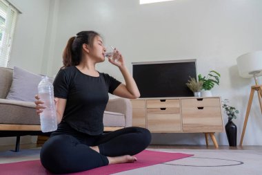 Çağdaş bir oturma odasında yoga minderinin üzerinde otururken siyah spor giysili bir kadın su içiyor..