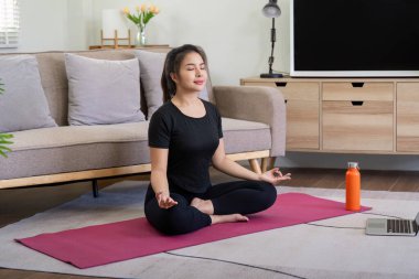 Siyah spor giysili genç bir kadın oturma odasında pembe yoga minderi üzerinde meditasyon yapıyor. Yanında bir laptop ve su şişesi var..