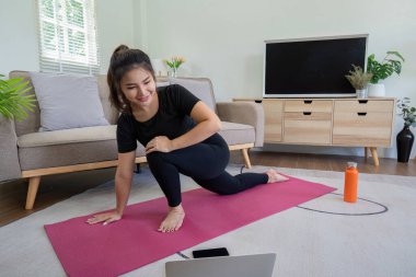 Siyah giysili bir kadın rahat, modern bir oturma odasında pembe bir minder üzerinde yoga yapıyor. Televizyon, bitkiler ve ahşap mobilyalar var..