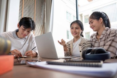 Üç öğrenci, parlak ve modern bir sınıf ortamında bir dizüstü bilgisayar ve kitap kullanarak bir projede işbirliği yapıyorlar..