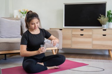 Siyah giysili genç bir kadın evde yoga yapıyor, pembe bir paspasın üzerinde oturuyor ve rahat bir oturma odasında bardağa su döküyor..