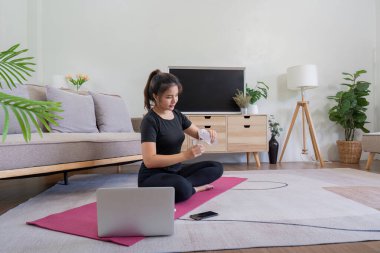 Evde yoga yapan genç bir kadın, rehberlik için dizüstü bilgisayar kullanıyor. Modern oturma odası dekorasyonu, kapalı mekan fitness ve sağlıklı bir yaşam tarzı..
