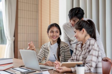 Modern bir çalışma odasında dizüstü bilgisayar kullanarak bir proje üzerinde çalışan üç öğrenci. Ortak öğrenme ve takım çalışması.