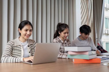 Modern bir sınıfta dizüstü bilgisayarlar ve kitaplarla birlikte okuyan üç öğrenci, takım çalışması ve odaklanma sergiliyorlar..