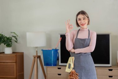 Önlüklü mutlu kadın, tamam işareti veriyor, modern oturma odasında temizlik malzemeleriyle duruyor, ev temizliğine hazır..