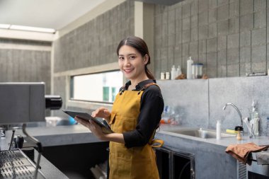 Önlük giyen genç bir barista, modern bir kahve dükkanında elinde tabletle kameraya gülümsüyor..