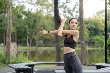 Atletik giyinen genç bir kadın parkta esneyerek dışarıda antrenman yapmaya hazırlanıyor. Etrafı yeşilliklerle çevrili..