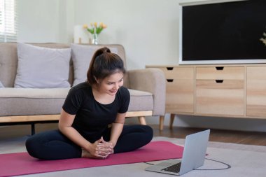 Aktif giyimli genç bir kadın evinde yoga yapıyor, rahat bir oturma odasında bilgisayarındaki sanal fitness dersini takip ediyor..