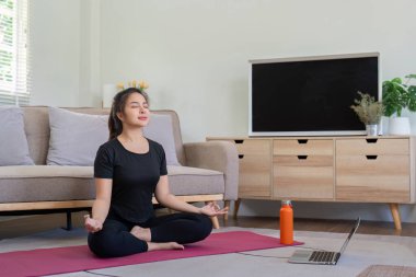 Siyah kıyafetli genç bir kadın, modern bir oturma odasında yoga ve meditasyon yapıyor. Rehberlik için dizüstü bilgisayar kullanıyor..