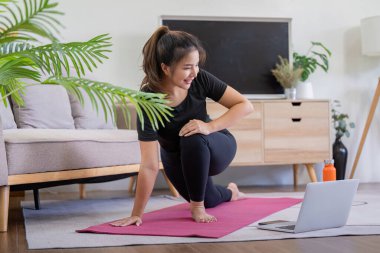 Pembe bir minderde yoga yapan bir kadın, rahat, modern bir oturma odasında bilgisayarındaki bir dersi takip ediyor bitkiler ve şık mobilyalarla..