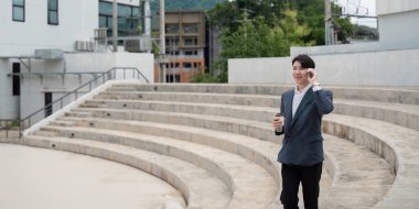 Takım elbiseli genç bir iş adamı açık hava amfitiyatrosunda dururken telefonla konuşuyor ve elinde kahve fincanı tutuyor..