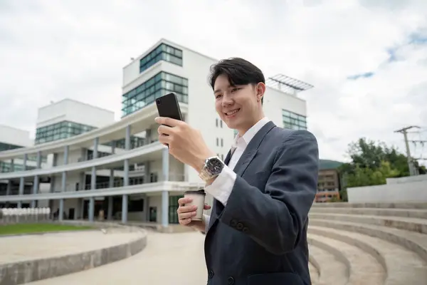 stock image Smiling young businessman in a suit using a smartphone while holding a coffee cup, standing outside a contemporary office building.