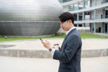 Takım elbiseli genç işadamı elinde kahve fincanı tutarken akıllı telefon kullanıyor, modern bir binanın önünde duruyor..