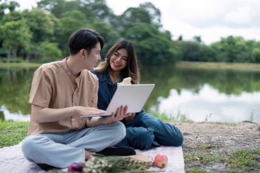 Gölün kenarında battaniyeye oturmuş, pikniğin tadını çıkaran ve doğayla iç içe dizüstü bilgisayar kullanan genç bir çift..