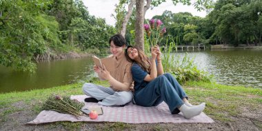 Sırt sırta verip göl kenarında piknik yapan neşeli bir çift, doğada huzurlu bir günün tadını çıkarıyorlar..