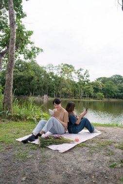Genç bir çift gölün kenarında huzurlu bir anın tadını çıkarıyor, yemyeşil bir parkta piknik battaniyesinde kitap okuyor ve dinleniyorlar..
