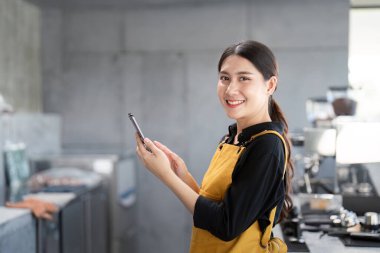 Sarı önlük giyen genç bir bayan barista modern bir kahve dükkanında akıllı telefon kullanırken gülümsüyor..