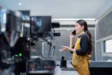 Sarı önlük giyen genç bir barista, modern bir kahve dükkanında tezgahın arkasında çalışırken telefonla görüşüyor..