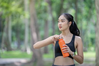 Spor kıyafetli genç bir kadın parkta elinde turuncu bir su şişesi tutuyor. Formu, su ihtiyacını ve sağlıklı bir yaşam tarzını temsil ediyor..