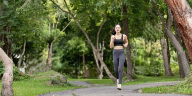 Atletik giyinen bir kadın, yemyeşil ağaçlar ve bitkilerle çevrili bir yolda koşuyor, fitness ve sağlıklı bir yaşam tarzını destekliyor..