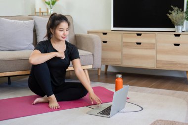 Siyah spor kıyafetli genç bir kadın evde laptopla yoga yapıyor, modern bir oturma odasında pembe bir paspasın üzerinde oturuyor..