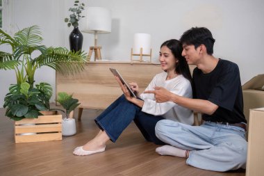 Neşeli bir çift yeni evlerinin zemininde oturuyor, iç tasarımlarını planlamak için tablet kullanıyorlar, kutular ve ev bitkileriyle çevrili..