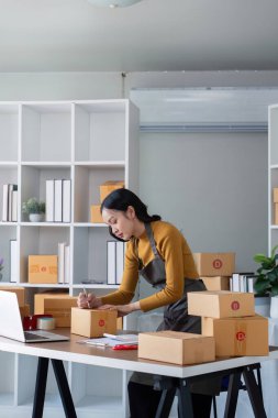 Modern bir ofiste nakliye için kutuları toplayan genç bir kadın bir dizüstü bilgisayarla masa başında çalışıyor ve paketleri düzenliyor..