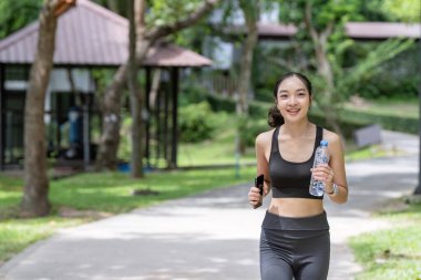 Parkta koşan genç bir kadın elinde su şişesi ve akıllı telefonla güneşli bir günün tadını çıkarıyor ve formda kalıyor..