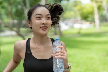 Gülümseyen genç bir kadın parkta koşuyor, elinde bir su şişesi tutuyor, güneşli bir günün tadını çıkarıyor ve aktif kalıyor..