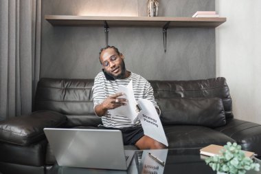 Genç Afrikalı Amerikalı adam evde birden fazla iş yapıyor, dizüstü bilgisayarda çalışıyor, belgeleri inceliyor ve telefonda konuşuyor..