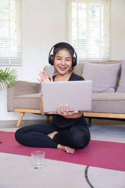 Oturma odasında yoga minderinin üzerinde otururken kulaklıkla gülümseyen genç bir kadın..